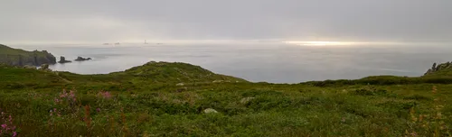 This panoramic landscape photograph captures the Cornish sun and mist during an early summer evening along the rugged coastline of Cornwall. The view showcases lush green cliffs descending gently towards the calm sea, with wildflowers in the foreground adding seasonal detail. Out to sea, the famous Longships Lighthouse is visible, rising from the mist as a distinctive landmark, surrounded by scattered rocks. The low sun is partially obscured by atmospheric mist, producing a soft glow on the water and enhancing the atmospheric quality of the Cornish coastal scene.